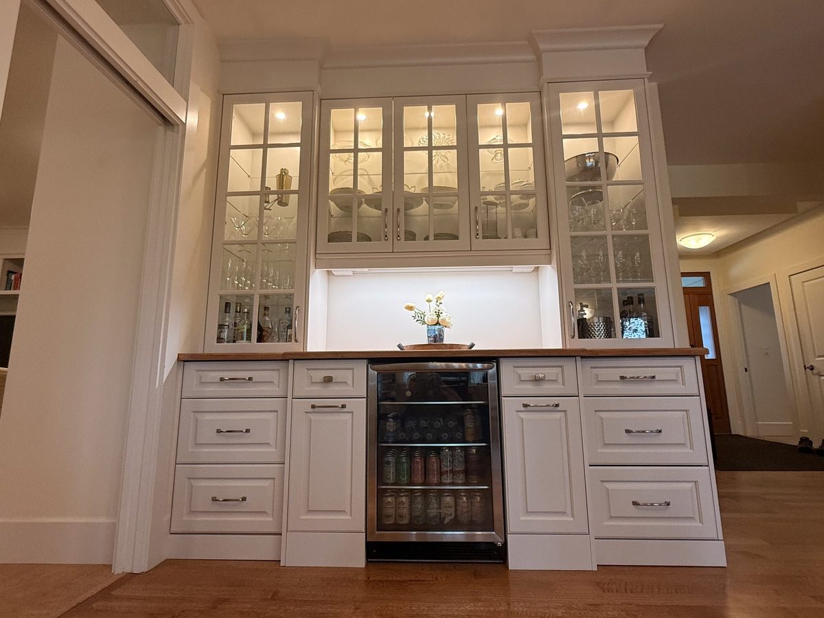 The Pouring Perfection dry bar in Nanaimo - lit glass-front upper cabinets over white raised-panel drawers, a wood counter and a built-in beverage fridge