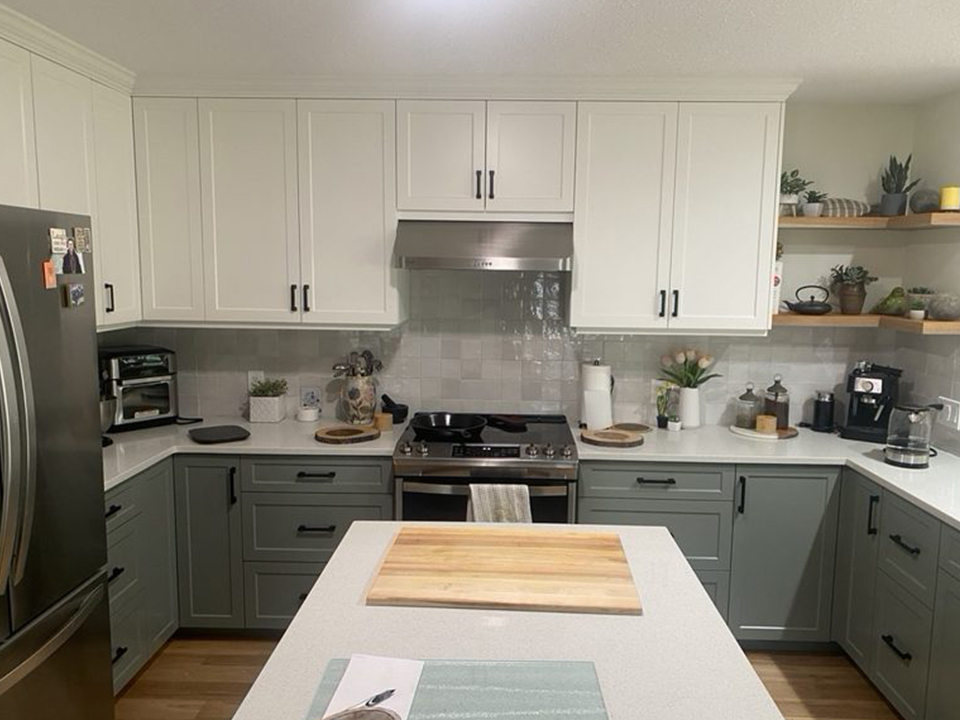 A two-tone kitchen with white upper cabinets, sage-grey lowers, quartz counters and floating wood shelves