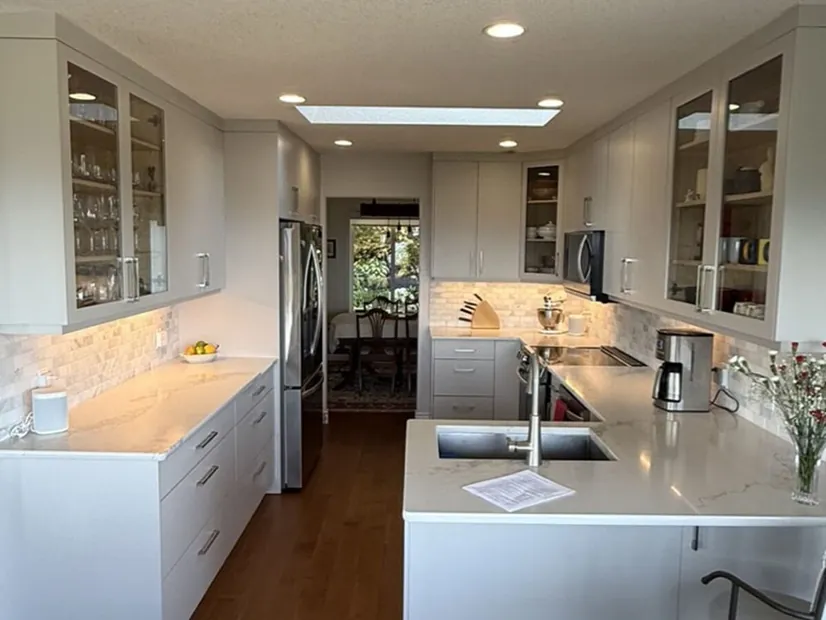 A Nanaimo galley kitchen with light grey cabinets, glass-front display uppers, marble-look backsplash and a quartz peninsula